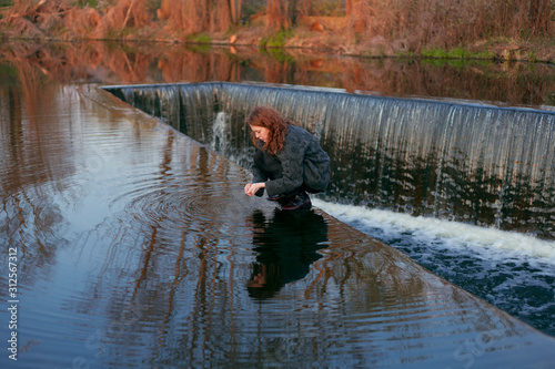 beautiful girl with red hair stands in the water at the edge of the waterfall washes her hands photo