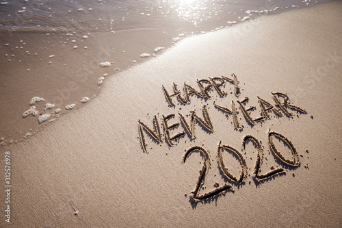 Happy New Year 2020 message handwritten in raised textured letters on a sandy beach with copy space next to incoming wave