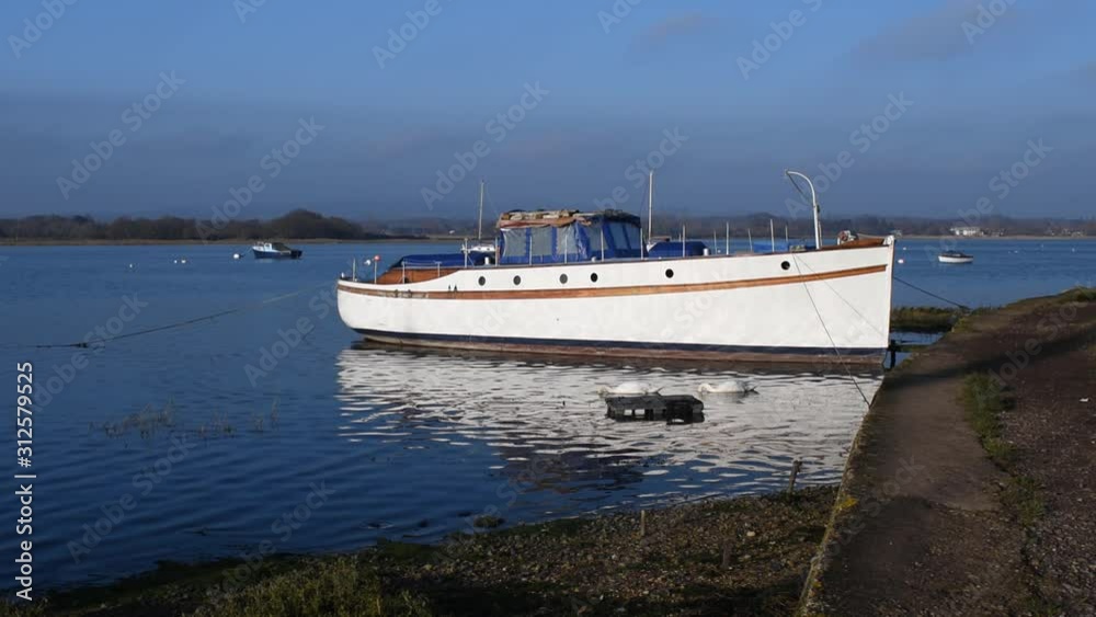Dell Quay near Chichester, a view of the inlet on a calm and clear winter day, a beautiful old boat reflecting in the calm water with a pair of swans in the foreground. Dell Quay near Chichester, a view of the inlet on a calm and clear winter day, a beautiful old boat reflecting in the calm water with a pair of swans in the foreground.