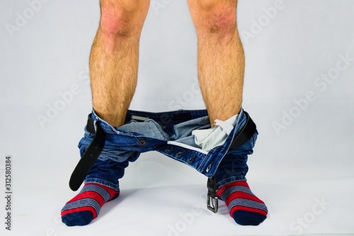 Man has his pants and underwear down  at the ankles.Man in front of white background has pants pulled down.