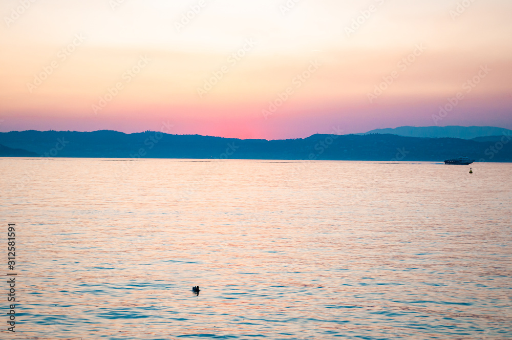 Naklejka premium Sunset above Garda lake with floating ships, freshwater birds resting on water surface and dolomite mountains on the horizon. Lombardy, Italy