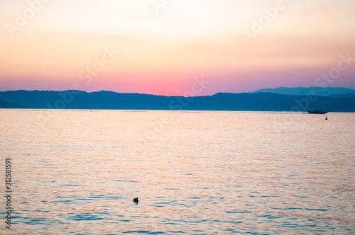 Sunset above Garda lake with floating ships  freshwater birds resting on water surface and dolomite mountains on the horizon. Lombardy  Italy
