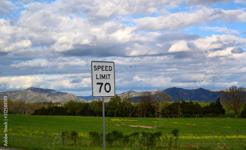 a 70 mph speed limit sign stands in the foreground with a beautiful sky and mountain range in the background on a sunny spring afternoon