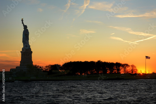 Statue of Liberty in silhouette at sunset viewed from the water with Flag, clouds and colorful sky.