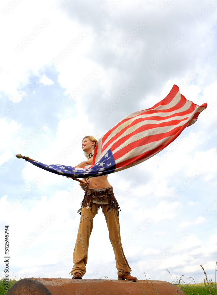 A beautiful young American girl stands on rusty cylinder. She proudly waves the US flag   cheerfully as the wind blows through her hair.