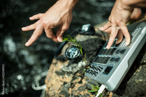 Obraz na plátně Caucasian male performing music using sampler in the nature sitting on a rock near the banks of crystal clear river
