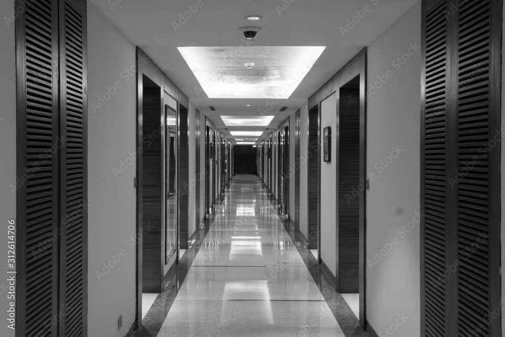 Horizontal black and white view of the brightly illuminated long tunnel shaped corridor with mirrors, wooden doors and cabinets on side walls