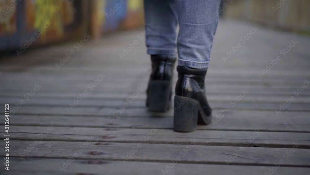 Close-up of women's legs in jeans and black heeled shoes, they walk on a wooden bridge, in the background a wall with graffiti. Close-up of women's legs in jeans and black heeled shoes, they walk on a wooden bridge, in the background a wall with graffiti.