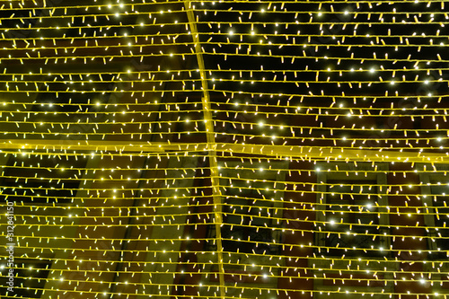 A festive garland with numerous yellow lights on a cross frame shines in the dark against the background of the building. Colorful and magical decoration for the holidays