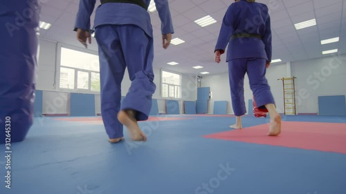 Following shot of barefoot male and female jujutsu athletes in blue uniform walking in martial arts gym, greeting each other with hand slap and start sparring fight