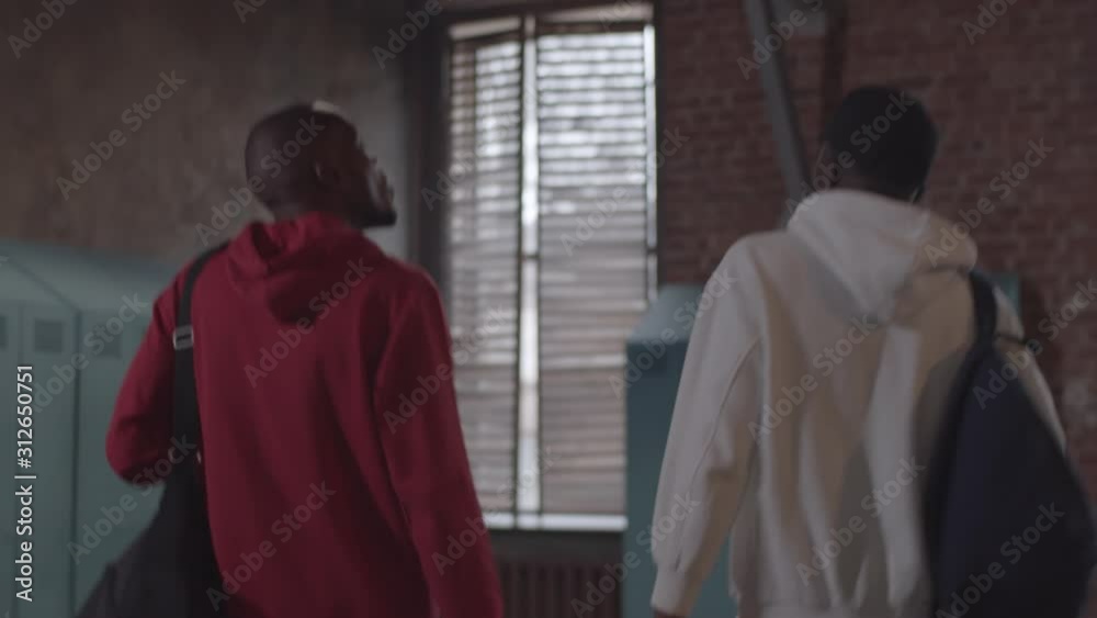 Waist-up rear tracking shot of two young African American sportsmen walking into changing room at gym, chatting, then bumping fists and putting down backpacks and bags next to lockers Waist-up rear tracking shot of two young African American sportsmen walking into changing room at gym, chatting, then bumping fists and putting down backpacks and bags next to lockers