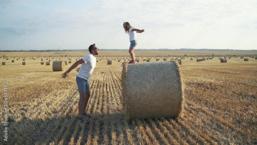 Little daughter jumping on father's arms from a big haycock in field Little daughter jumping on father's arms from a big haycock in field