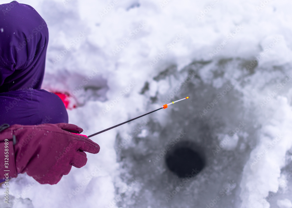 The hand of a gloved angler girl in winter holds a small fishing rod with a nod to a hole in the ice for the purpose of catching fish.
