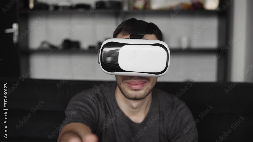 Young bearded man wearing headset of virtual reality in office sitting on a sofa and trying to touch something that he see in virtual world. Young bearded man wearing headset of virtual reality in office sitting on a sofa and trying to touch something that he see in virtual world.