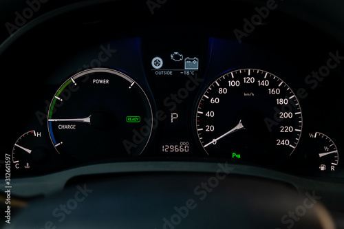 The dashboard of car is glowing white with arrows at night with a speedometer, charge battery level and other tools to monitor the condition of the hybrid vehicle in modern style on black background