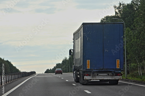 Big Semi-truck with blue semi trailer driving on suburban empty highway road on Summer day on green trees background, rear view © Ilya