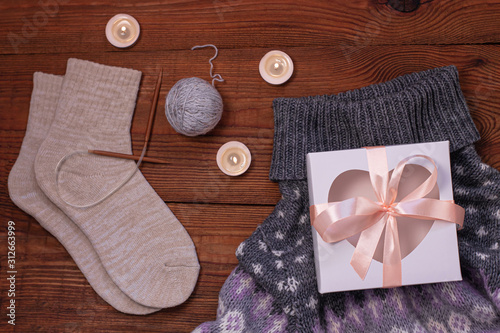 Cozy flatlay. Gift box, knitted socks, ornament sweater, candles, knitting needles, yarn on wooden background flat lay.Winter holidays concept.Warm weekend cold weather.Trendy knitted clothes,top view