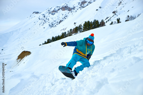 A young man in a colored winter hoodie and mask rides in the mountains on a snowboard in a cool hat with an orange pom pom