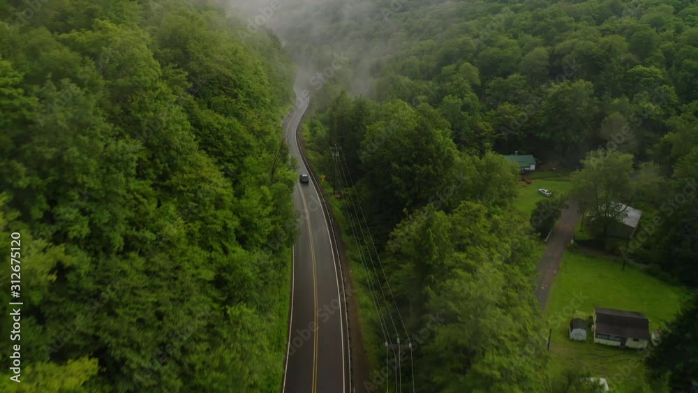 Aerial drone overhead of single car driving down curvy mountain road through green forest valley with trees and rainy fog
