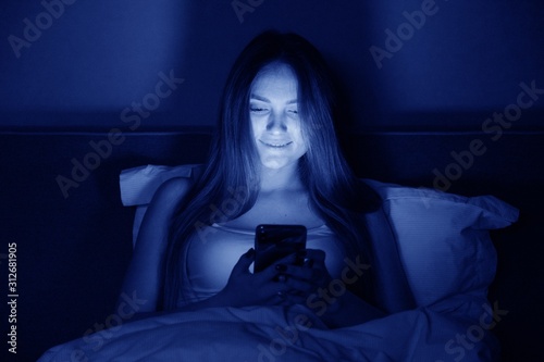 Young beautiful woman at night sitting in bed with smartphone