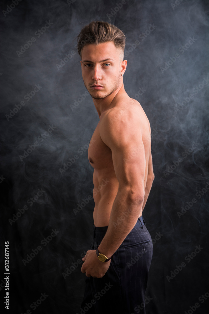 Handsome shirtless athletic young man wearing only pants, with blue eyes, looking at camera in studio shot, on dark background