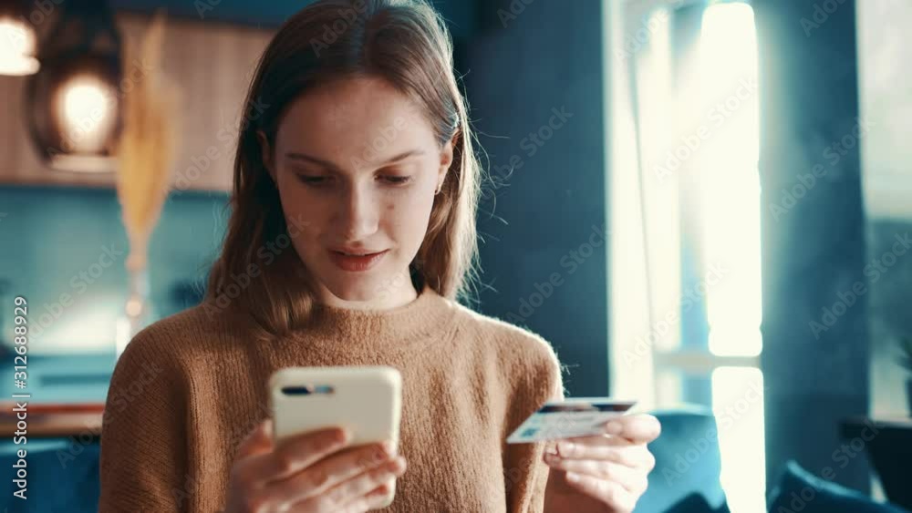 Cheerful redhair caucasian girl is making online payment holding bank card using modern smartphone or cell phone at home, sitting on sofa and smiling. Finance, shopping in internet.