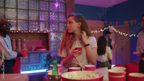 Upset attractive blonde girl using smartphone internet app getting bored jealous while staying at night party friends reunion. Concept of unsociable people.