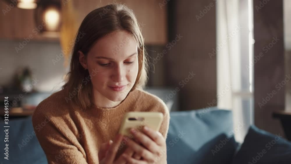 Attractive redhair teenager girl sitting of the sofa at home using phone and smiling. Feeling happy. Communication female looking message at cell phone or smartphone. Slow motion.