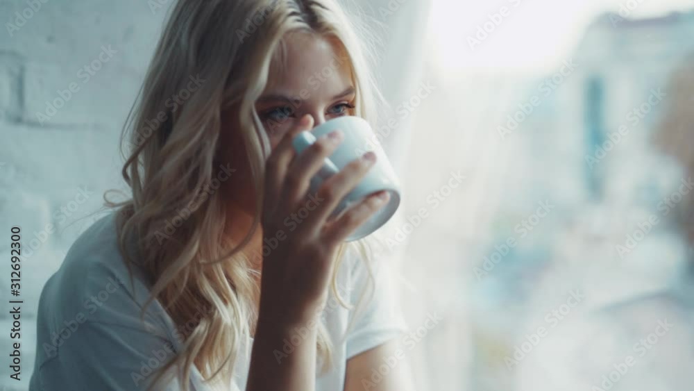 Happy beautiful woman smiling and drinking a cup of coffee or tea near the window relax in the morning cozy young home enjoying attractive comfortable relax warm slow motion