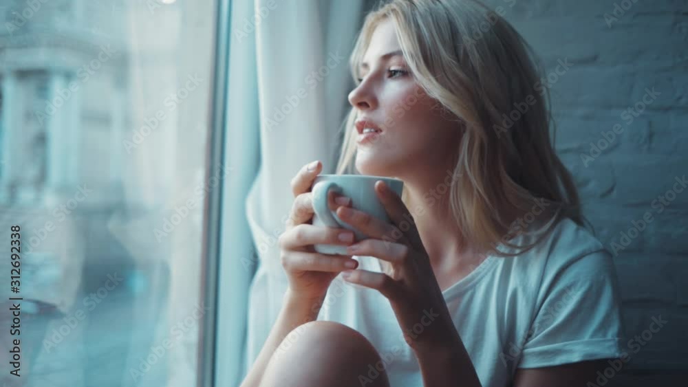 Smiling beautiful woman drinking a cup of coffee or tea near the window relax in the morning cozy young home enjoying attractive comfortable relax warm slow motion