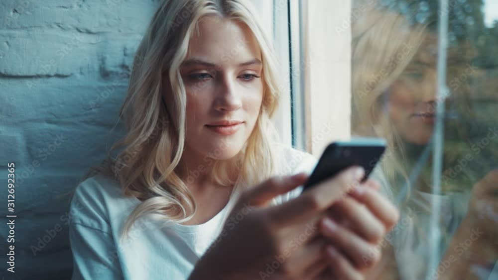 Close up young woman sitting on a windowsill use phone communication female message cellphone cheerful smile internet modern smartphone portrait slow motion Close up young woman sitting on a windowsill use phone communication female message cellphone cheerful smile internet modern smartphone portrait slow motion