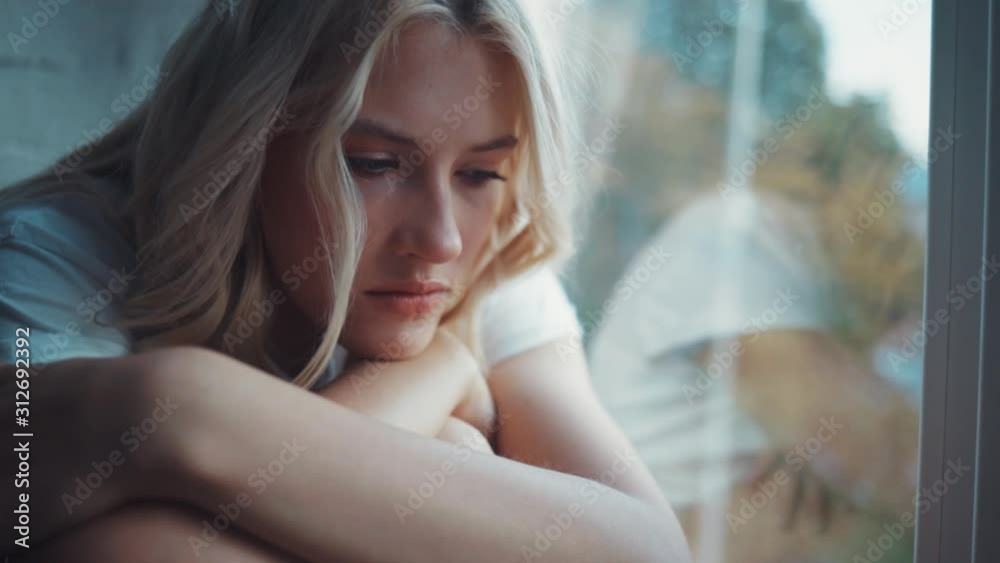 Sad woman sits on the windowsill looks out the window frustration sickness hopeless loneliness depression alone beautiful home bed lonely gown comfortable close up slow motion