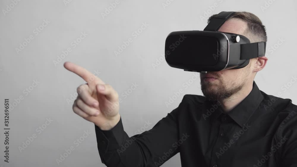 Young bearded man sitting against computer with virtual reality glasses trying to touch something or tap some thing in front of what he seeing. Modern technologies. The concept of future technology. Young bearded man sitting against computer with virtual reality glasses trying to touch something or tap some thing in front of what he seeing. Modern technologies. The concept of future technology.