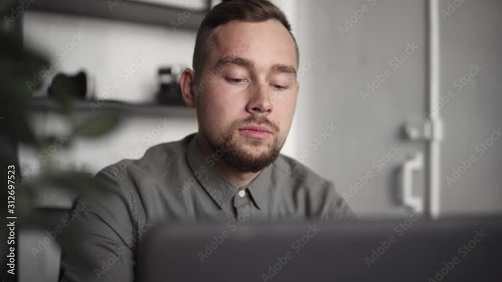 Young businessman or student in a shirt sitting against laptop. Working on a pc at a table in the office with a thoughtful expression. Young businessman working on his laptop in office. Young businessman or student in a shirt sitting against laptop. Working on a pc at a table in the office with a thoughtful expression. Young businessman working on his laptop in office.