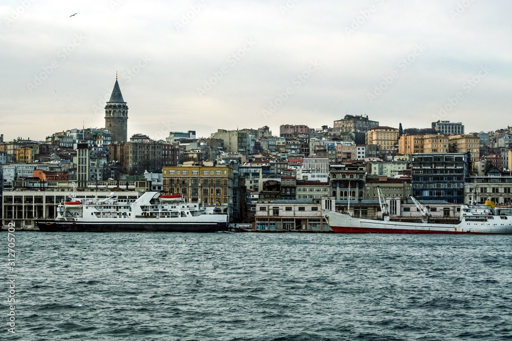 Galata Tower, on its hill, in Karakoy and Beyoglu district, taken during a cloudy winter afternoon, while the sea, ferry boats and cargo ships can be visible in foreground in Istanbul, Turkey