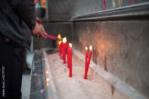 Group of red lighting candles put in the sand in concrete tray with blurred hands holding incenses