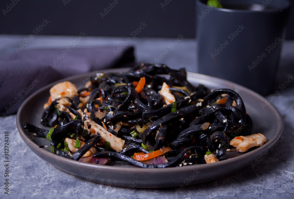 Black squid ink udon noodles with chicken meat on the black plate and cup of tea on dark stone background. Black squid ink udon noodles with chicken meat on the black plate and cup of tea on dark stone background.