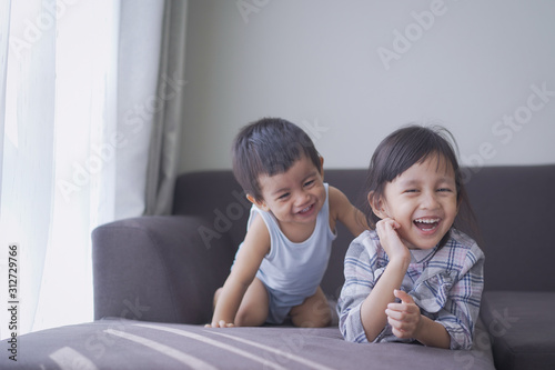Adorable asian little sister and her baby brother are playing and laughing together on the sofa in the morning time, concept of love and relation of child sibling in the asian family lifestyle. 