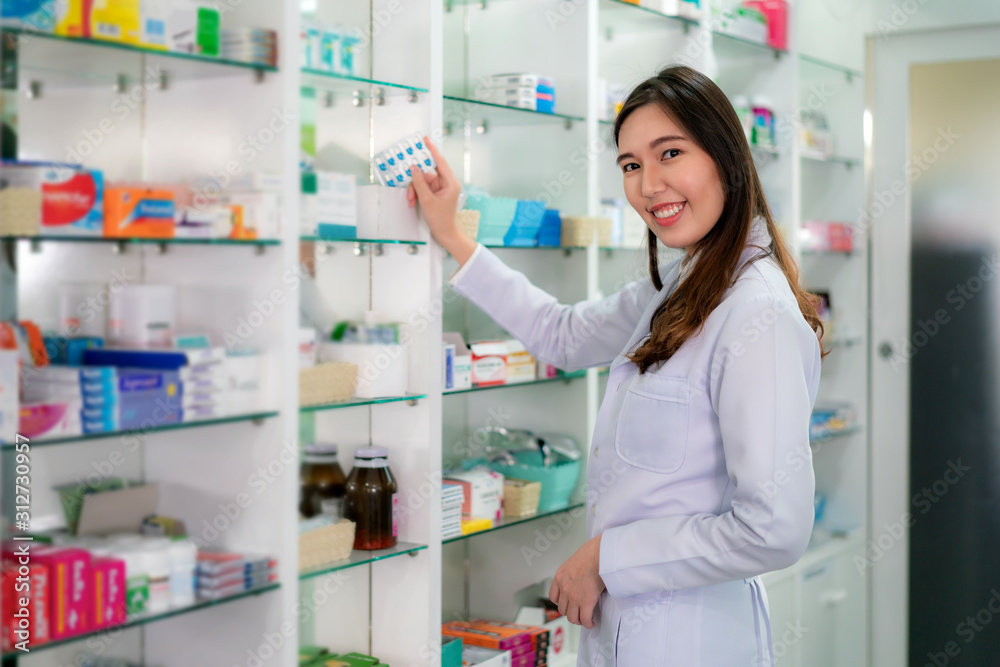 Asian young woman pharmacist with a lovely friendly smile pick capsule medicine and looking at camera in the pharmacy drugstore. Medicine, pharmaceutics, health care and people concept.