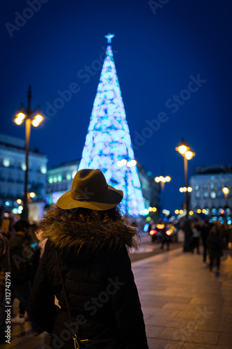 Tourist woman with warm clothes looking at Puerta del Sol square illuminated by christmas lights and a shinny christmas tree.