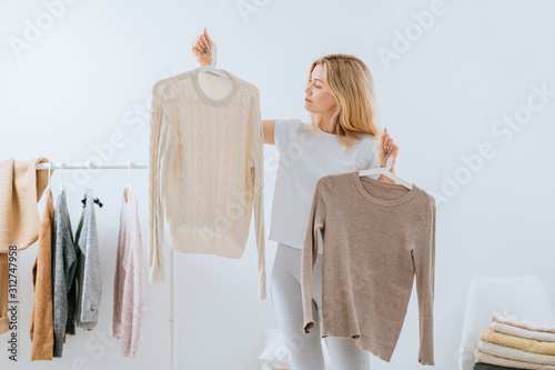Young pretty girl blogger stands with a thoughtful expression on his face, having difficult choice not knowing what to put on on the background of clothes hanging on a hanger in the wardrobe.