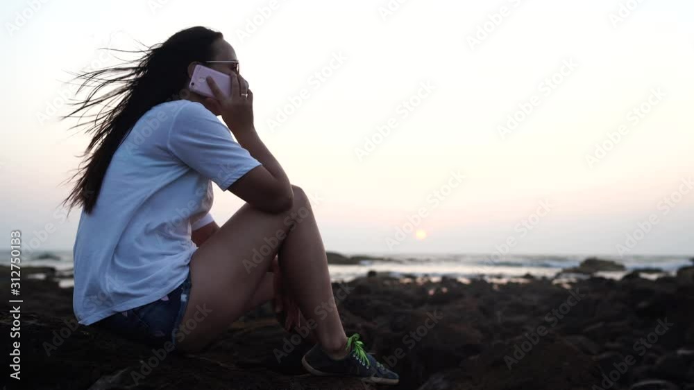 Relaxed woman sitting on stone and speaking on smartphone on shore. Side view of pleasant woman spending time enjoying vacation and talking on mobile phone on stony shore Relaxed woman sitting on stone and speaking on smartphone on shore. Side view of pleasant woman spending time enjoying vacation and talking on mobile phone on stony shore