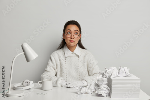 Pensive young female secretary wears big round glasses, white knitted jumper and gloves, looks aside, thinks deeply about something, has much crumpled papers, headphones and desklamp on desktop photo