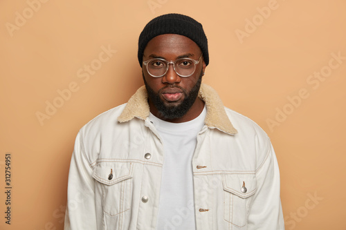 Studio shot of handsome dark skinned man with thick bristle wears transparent glasses, white shirt, black hat looks seriously at camera, being hard to impress, has calm expression, poses on beige wall