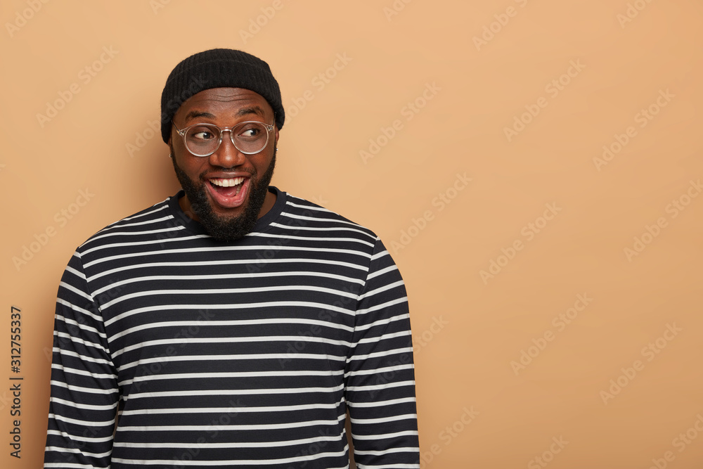 Half length shot of cheerful thoughtful adult man with dark skin, thick beard, opens mouth and concentrated aside, wears black hat and striped jumper, isolated over beige background, blank space Half length shot of cheerful thoughtful adult man with dark skin, thick beard, opens mouth and concentrated aside, wears black hat and striped jumper, isolated over beige background, blank space