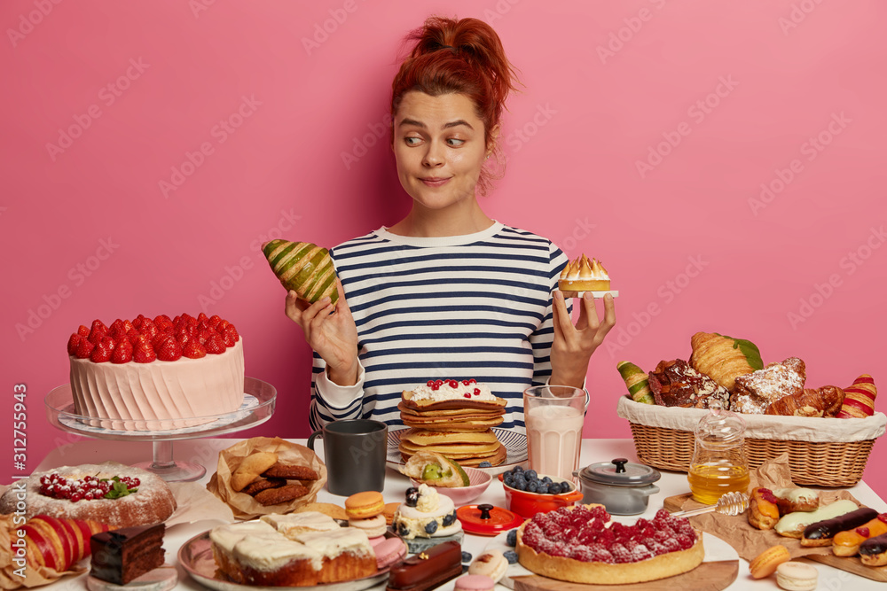 Pretty redhead woman chooses between croissant and cake, sits at table overloaded with desserts, dressed in striped jumper, isolated over pink wall. Unhealthy eating, harmful food and sugar obsession Pretty redhead woman chooses between croissant and cake, sits at table overloaded with desserts, dressed in striped jumper, isolated over pink wall. Unhealthy eating, harmful food and sugar obsession
