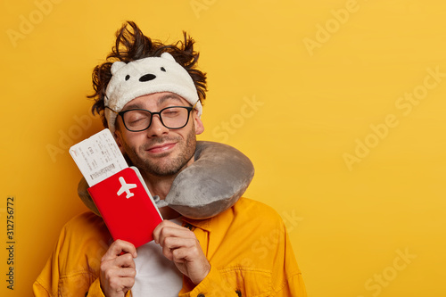 Pleased male passenger dreams about awesome trip abroad holds passport with flying tickets, stands with eyes shut, wears sleep mask and neck pillow for comfort travel, isolated on yellow wall photo