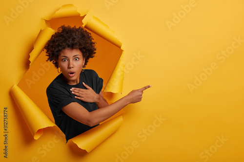 Stunned Afro American woman looks with scared speechless expression, holds breath from excitement, demonstrates something on right side, stands in paper hole, wears black t shirt advertises copy space
