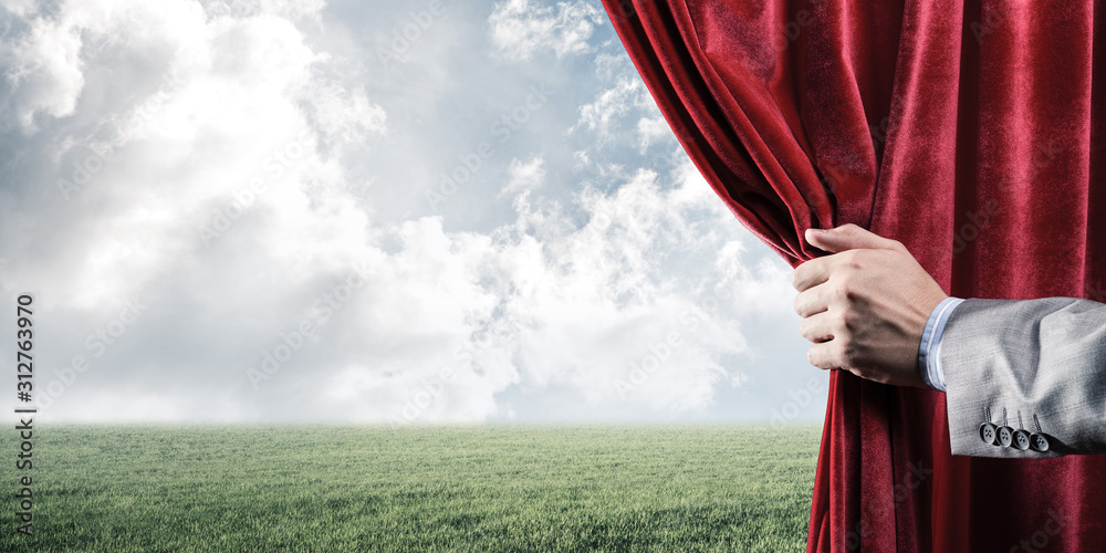 Fototapeta premium Green meadow behind red curtain and hand holding it
