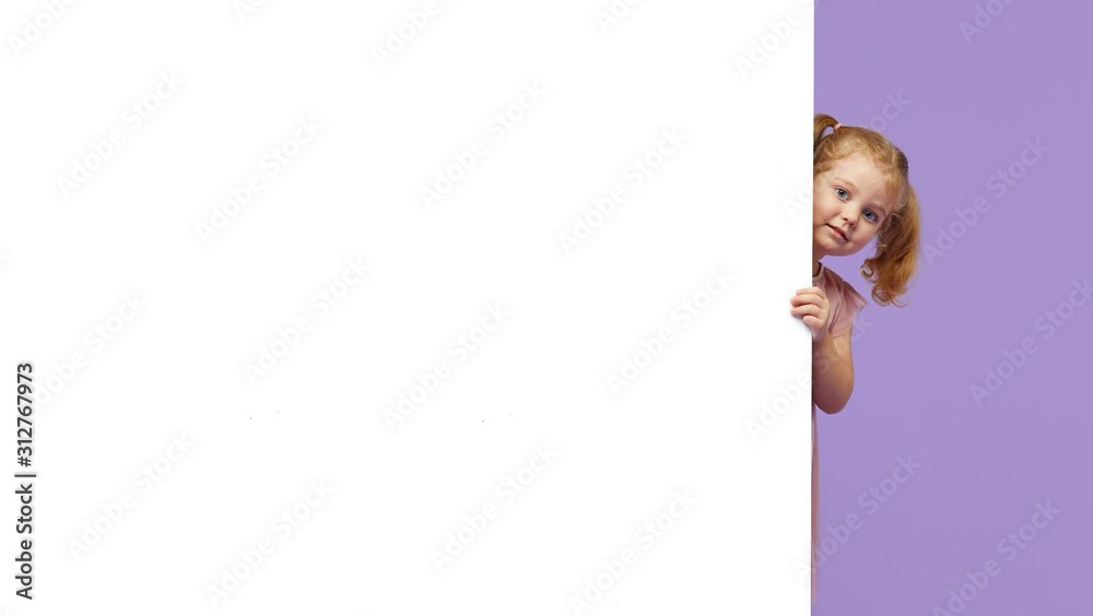 Little smiling child standing behind a white blank panel isolated against purple background. Peeking out from behind a banner, empty space for text. Little smiling child standing behind a white blank panel isolated against purple background. Peeking out from behind a banner, empty space for text.